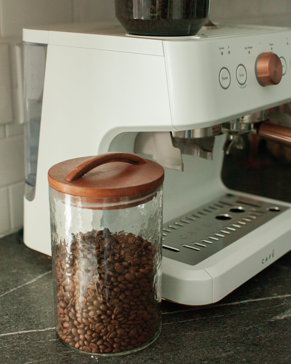 Clear glass canister with a wooden lid storing coffee beans beside an espresso machine