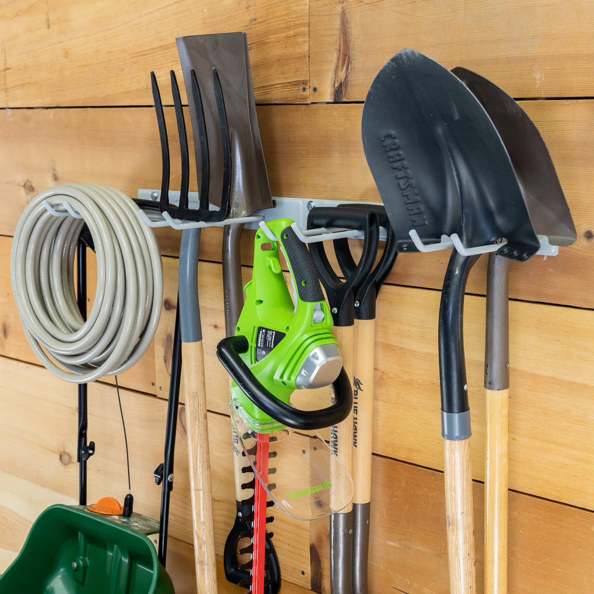 Gray garden tool organizer on a wood wall holding a hose, rakes, shovels and a trimmer