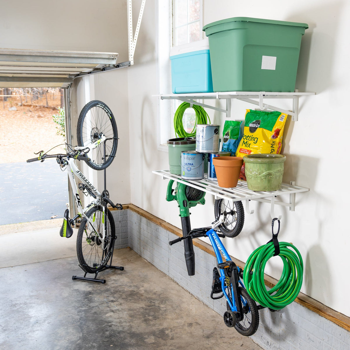 White wall shelves in a garage holding bins and garden supplies, with a bike and hose hung below
