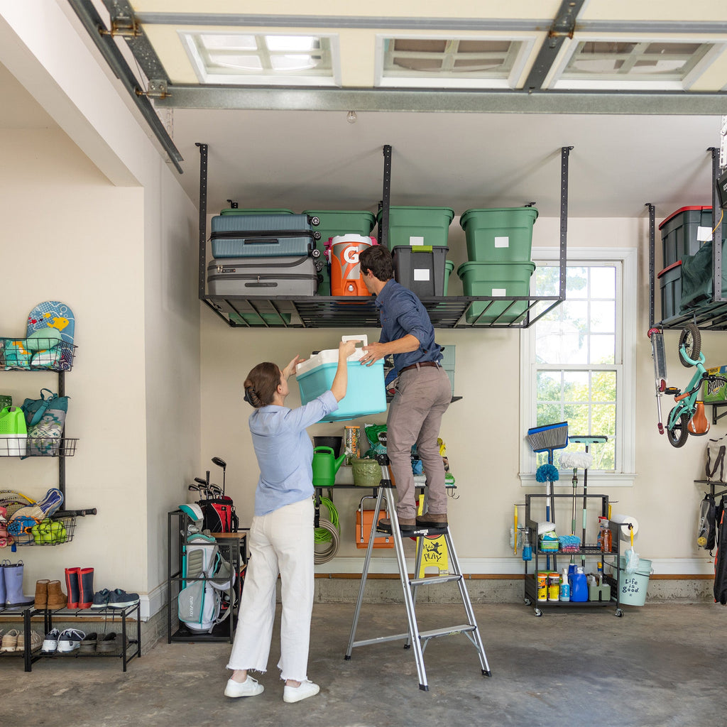 Two people loading bins onto overhead racks from a ladder in a large garage