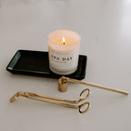 Matte black stoneware tray holding a lit candle beside a gold candle care kit
