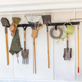 Two black garden tool organizer rails holding rakes, shovels, a hat, gloves and a hose in a shed