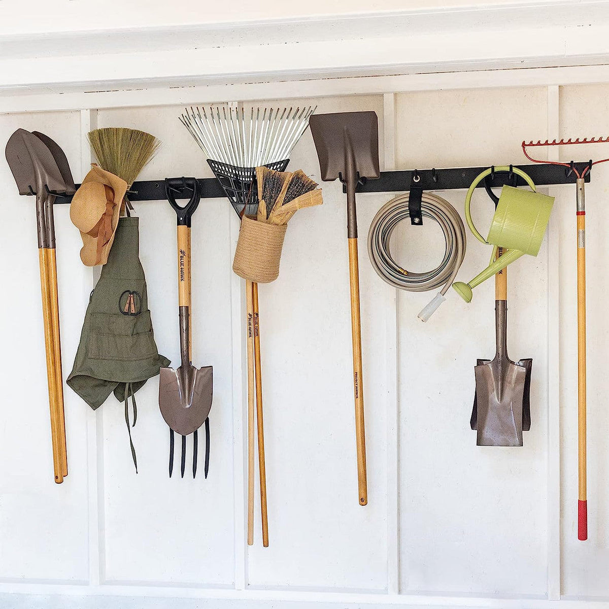 Two black garden tool organizer rails holding rakes, shovels, a hat, gloves and a hose in a shed