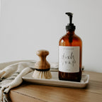 Cream stoneware tray holding an amber dish soap dispenser and a dish brush