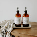 Cream stoneware tray holding two amber soap dispensers on a wooden counter