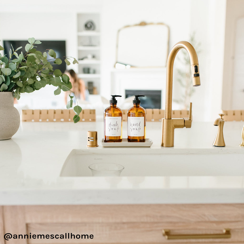 Cream stoneware tray with amber soap dispensers beside a kitchen faucet