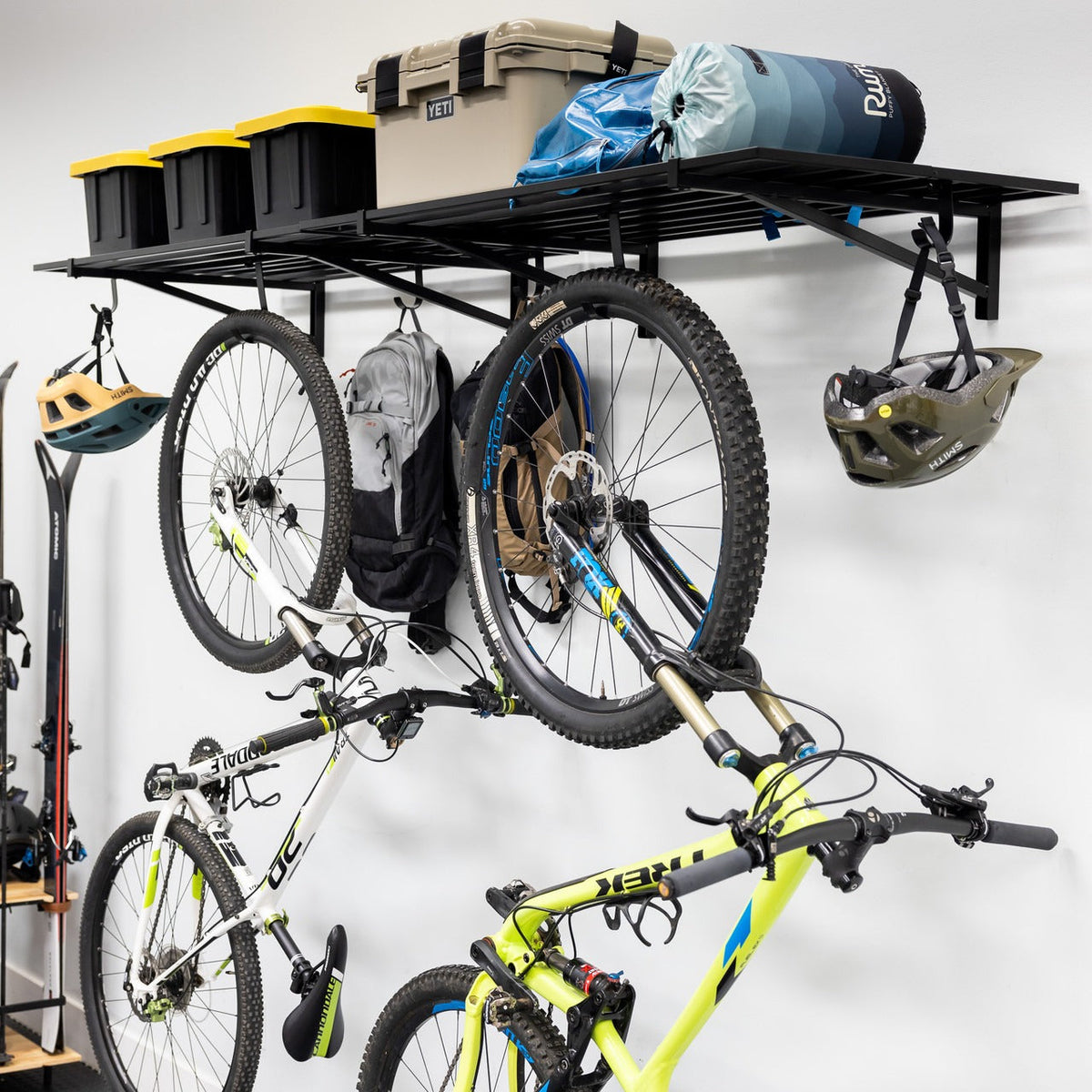 Two bikes hanging from hooks under a black wall shelf holding bins and a helmet