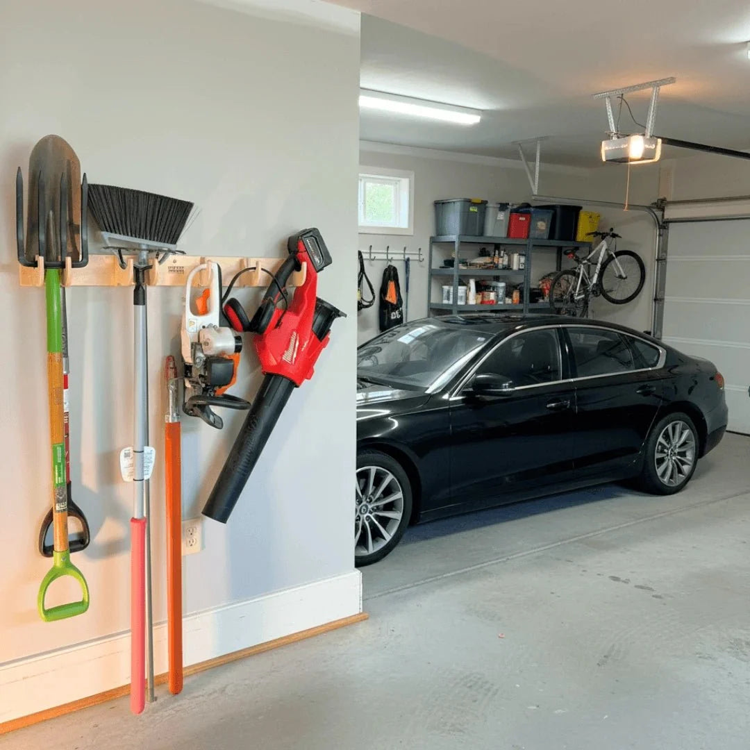 Hook rack holding yard tools and a leaf blower on a garage wall beside a car