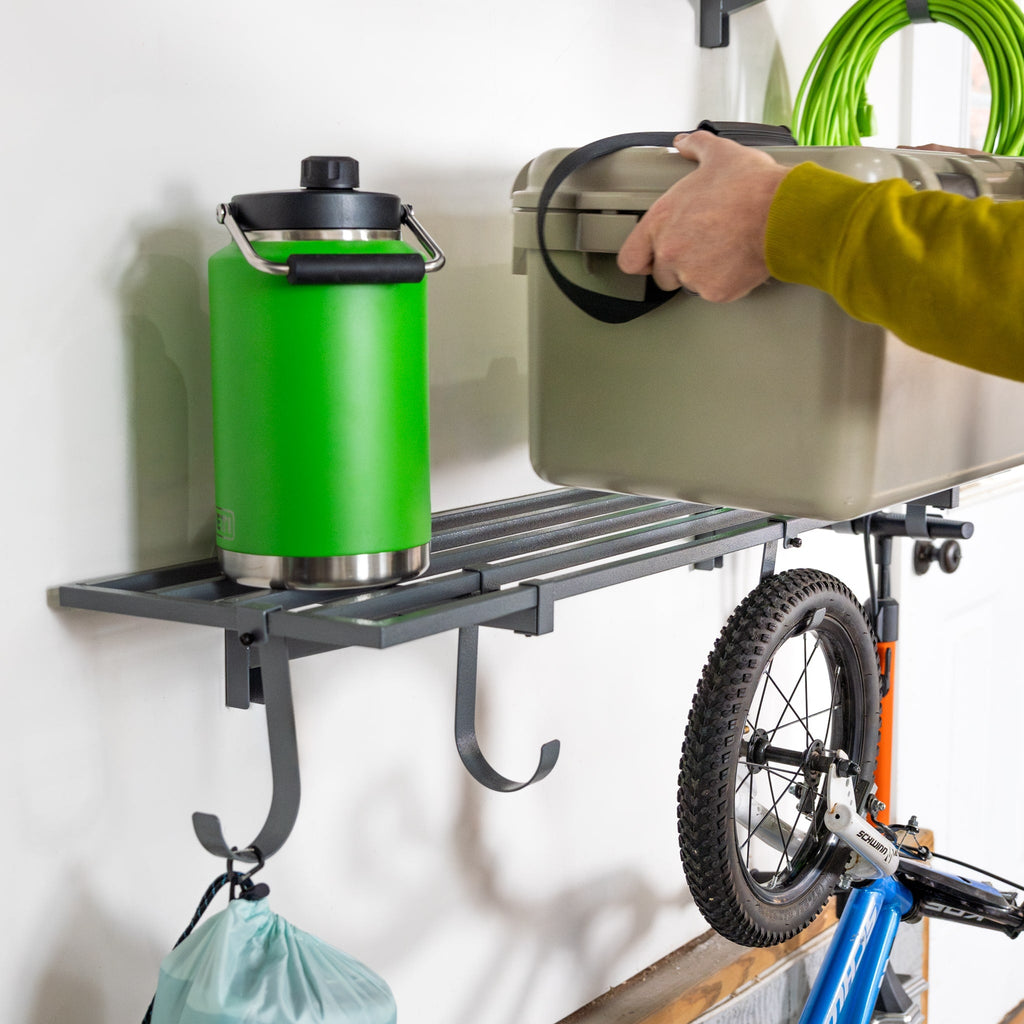 Hand placing a cooler on a gray wall shelf holding a water jug, with a bike hung below