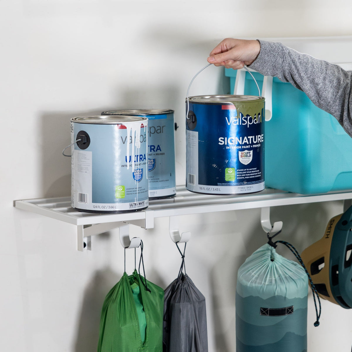 Hand placing a paint can on a white wall shelf with bags hung from the hooks below