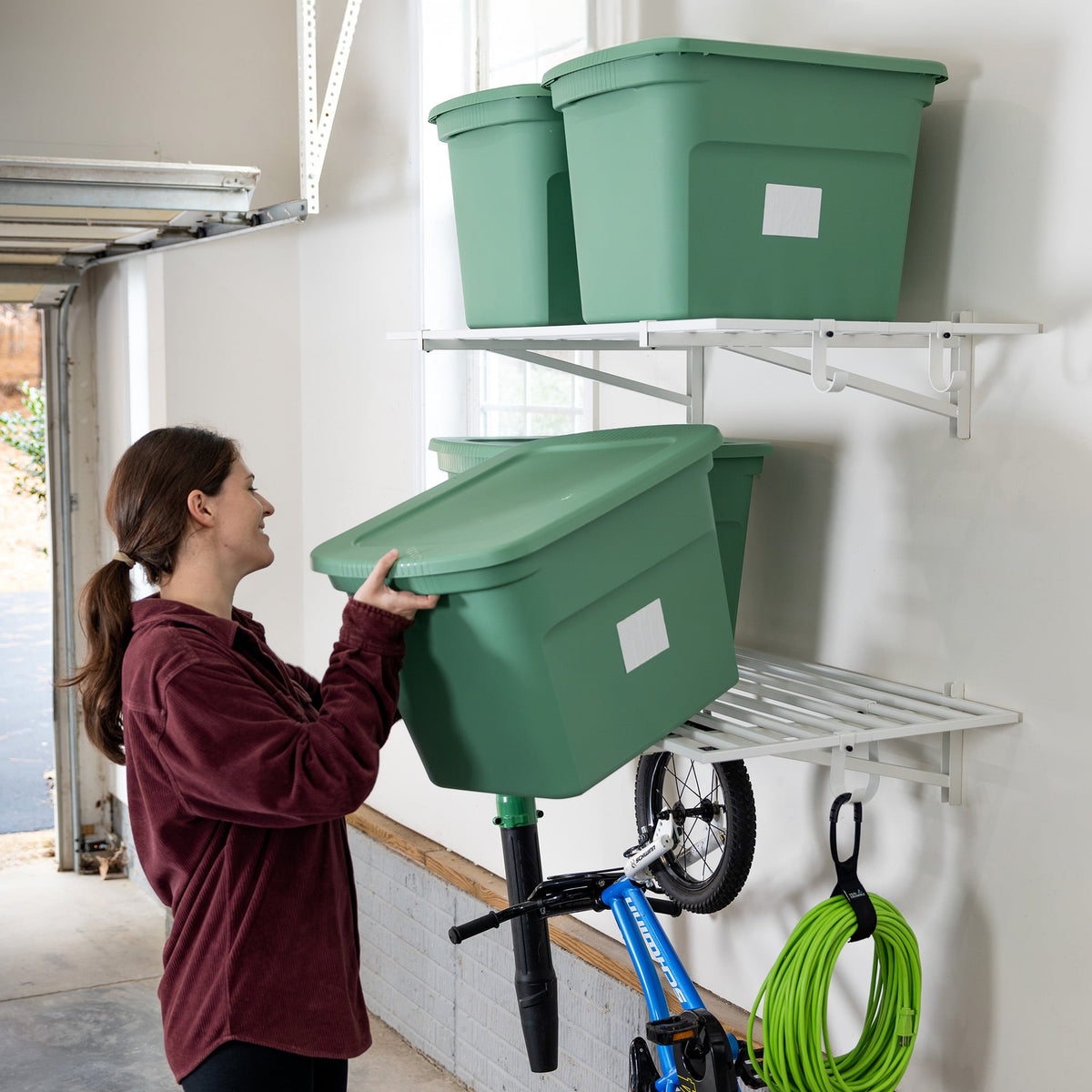 Person lifting a green storage bin onto a white garage wall shelf